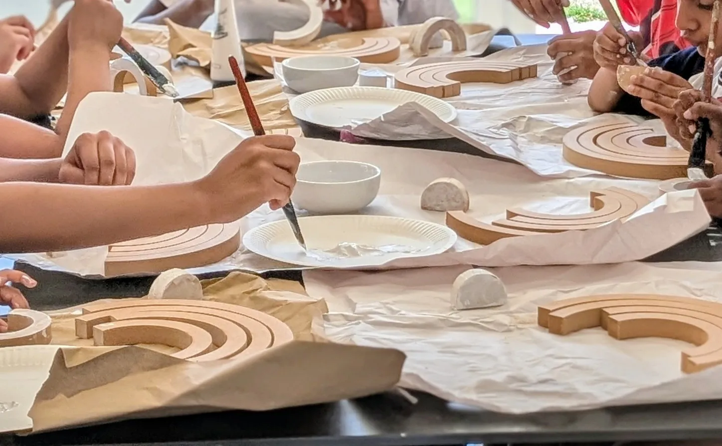 Children painting layered wooden rainbow shapes around a paper-covered studio table